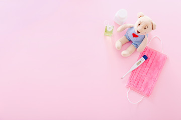 Protective medical mask with a bear toy, thermometer, and hand sanitizer on a pink background....