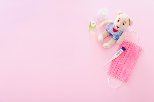 Protective medical mask with a bear toy, thermometer, and hand sanitizer on a pink background. Concept of quarantine of schools, home education