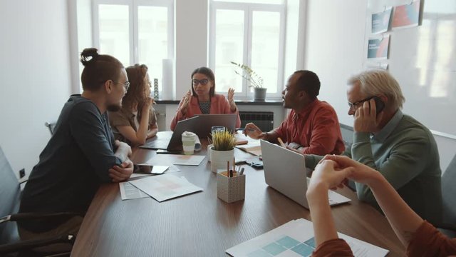 Zoom in shot of beautiful mixed raced female CEO talking to team of multiethnic colleagues while senior man having mobile phone conversation during meeting in office