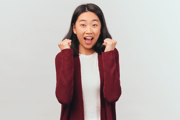 Enthusiastic girl clenches fists, exclaims Yes, satisfactory result. Beautiful woman of Asian appearance dressed in burgundy cardigan stands isolated white background Studio