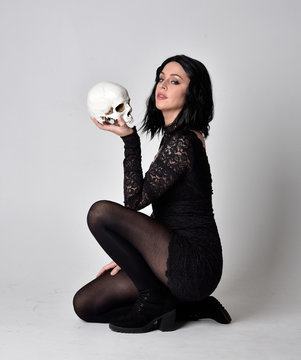 Portrait of a goth girl with dark hair wearing black lace dress and boots. Full length sitting pose and holding a human skull, on a studio background.