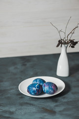  Happy Easter. Easter still life with colored eggs, a white plate and a white vase with dry twigs on a blue table.