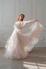 Beautiful natural redhead girl bride, with nude makeup, wearing a white dress, sensual dances in light interior.
