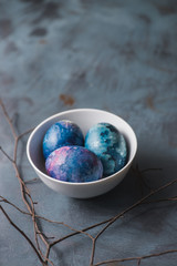 
Easter colorful eggs in a white bowl and branch on a blue textured background.
