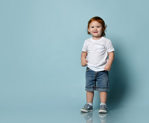 Little ginger toddler male or female in white t-shirt, socks and shoes, denim shorts. Child is...