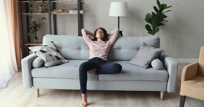 Calm young adult lady lounging on comfortable sofa in modern living room interior. Relaxed tired woman napping on couch hands behind head. Smiling girl feels no stress enjoying peaceful chill at home.