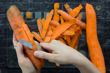 Top view of woman hands peeling carrot with knife on glass cutting board on the black background
