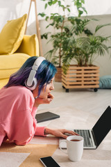Freelancer with colorful hair and headphones working with laptop near cup of tea, notebook and smartphone on floor