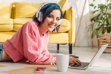 Fototapeta na wymiar Freelancer with colorful hair in headphones working with laptop near cup of tea and smartphone, smiling and looking at camera on floor