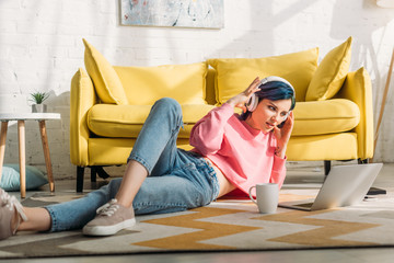 Freelancer with colorful hair and headphones looking at laptop and lying near sofa and cup of tea...