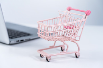 Online shopping. Mini empty pink shop cart trolley over a laptop computer on white table background, buying at home concept, close up