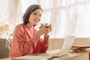Freelancer with colorful hair, cup of tea and pen looking at camera and smiling at table with laptop in living room