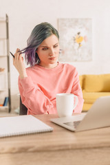 Selective focus of thoughtful freelancer with colorful hair holding pen at table with laptop, cup of tea and notebook in living room