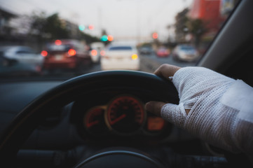 Closeup of injured hand with white bandage holding steering wheel with blurred cars background,...