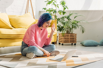 Girl with colorful hair waving hand, smiling and making video call on floor in living room