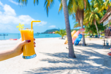 First-person view. Girl holds a glass cup of cold mango fresh on the background of a sandy tropical beach. White sand and palm trees. Fairytale vacation