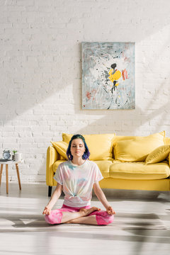 Girl with colorful hair and closed eyes meditating on yoga mat in living room