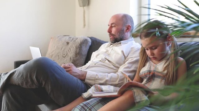 Father using laptop and daughter painting, sitting on couch at home