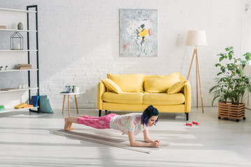 Obraz na płótnie Canvas Girl with colorful hair doing plank on yoga mat near sofa in living room