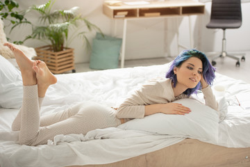 Beautiful woman with colorful hair smiling and resting with crossed legs on bed