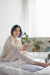 Freelancer with colorful hair smiling, looking at camera and using smartphone near laptop, notebook and cup of tea on bed