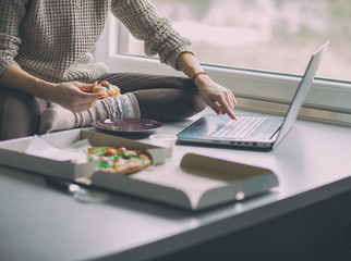 Young woman with laptop and pizza sitting home by the window, social media or home office concept