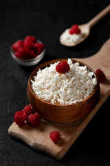 fresh homemade cottage cheese in a wooden bowl with raspberries on a dark background, healthy food on a black table