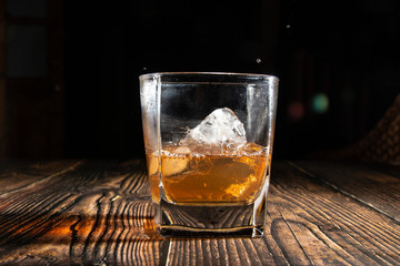 Whiskey with ice cubes on a wooden table. An old tabletop with light and a glass of strong drink. A splash of whiskey in a glass while serving ice.