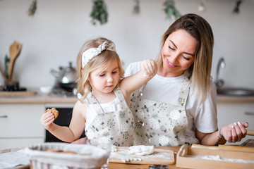 Mother and daughter prepare cookies in the kitchen at the table. A little blonde girl and a woman with light hair knead the dough and sprinkle it with flour.
