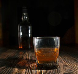 Glass of whiskey with ice cubes, a bottle on a wooden table. An old tabletop with light and a glass of strong drink.