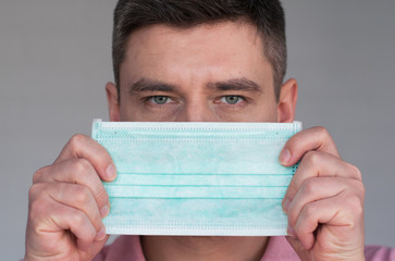 Portrait of dark hair european man holding surgery face mask in hands on light gray background