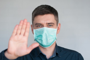 Portrait of european dark hair man in surgery face mask in blue shirt with stop palm on gray background