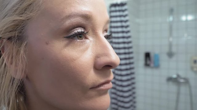 A young blond woman combs her eyebrows in front of a bathroom mirror.
