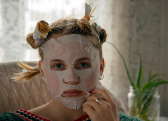 A teenage girl with curlers on her head and a cosmetic mask on her face against a background of lace curtains.