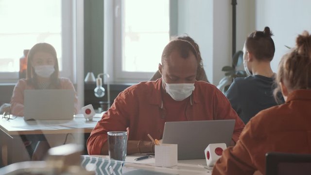 Young African American man in protective face mask sitting at table and using laptop while working in open space office with diverse colleagues during coronavirus outbreak
