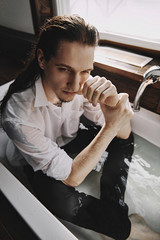 A guy with long black hair in jeans and a white shirt sits in a bathtub filled with water, next to the window