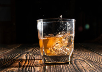 Whiskey with ice cubes on a wooden table. An old tabletop with light and a glass of strong drink. A splash of whiskey in a glass while serving ice.