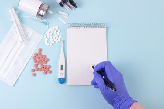 Medicine, Pharmacy, Health Concept. Doctor's Hand In Blue Medical Glove Write A Prescription. Blue Desk With Pills, Thermometr, Medicine Mask, Syringe. Mock Up Blank Empty Notepad. Top View Overhead.