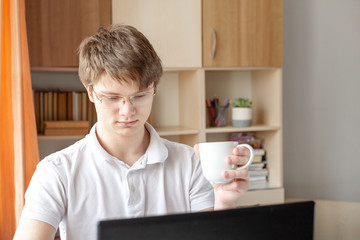 Student in glasses learning at home using laptop. Young man holding mug. Home school, online education, home education