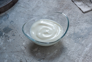 Natural yogurt in a transparent oval bowl standing on a concrete tabletop background. Copyspace, culinary cover.
