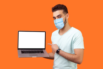 Portrait of positive joyful brunette man with surgical medical mask standing holding laptop with blank screen and smiling at camera, internet advertising. studio shot isolated on orange background