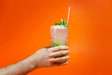 Close up of a man's hand holding a glass of tropical mojito cocktail on orange background with copy space. Summer time vacation concept.