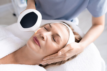 Above view of unrecognizable beautician using laser device while making skin rejuvenation procedure to mature woman