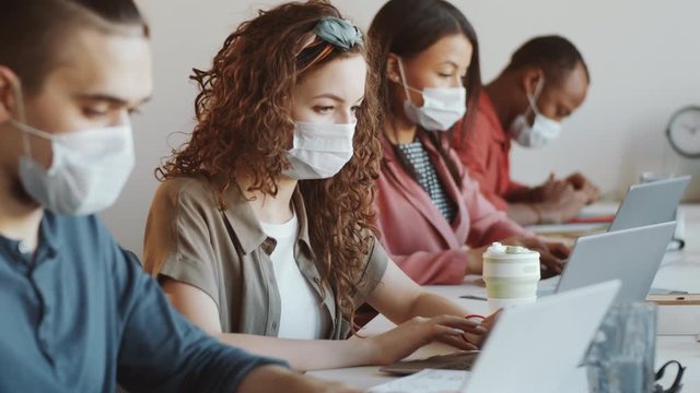 Rack focused shot of multiethnic young men and women in medical face masks working on laptops while sitting together at desk in open space office. Covid-19 outbreak concept