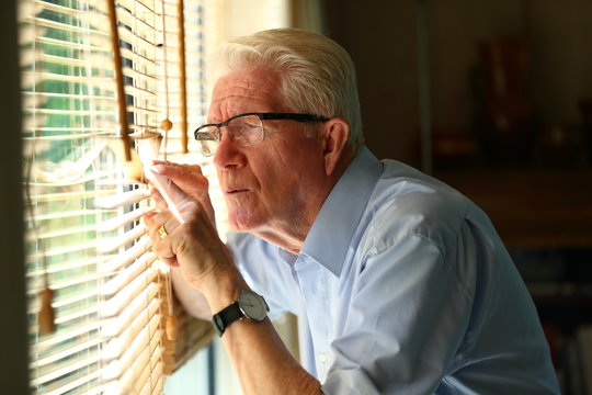 Curious elderly man peeking through the blinds by the window