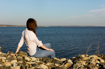 young longhaired woman in summer clothes sitting alone on the rocks at the beach and looking on horizon