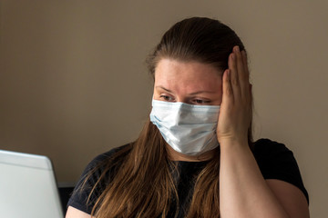 Girl in a medical mask looks at the laptop screen and reads the news about the coronavirus, grabs...