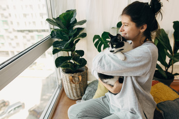 Happy hipster girl hugging cute cat, sitting together at home during coronavirus quarantine. Stay...