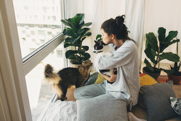 Hipster girl hugging and playing with two cats in modern room, sitting together at home during coronavirus quarantine. Stay home stay safe. Isolation at home to prevent virus epidemic.