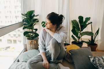 Sad hipster girl sitting at home during quarantine and looking at window. Stay home stay safe. Isolation at home to prevent virus epidemic. Young woman relaxing in modern room